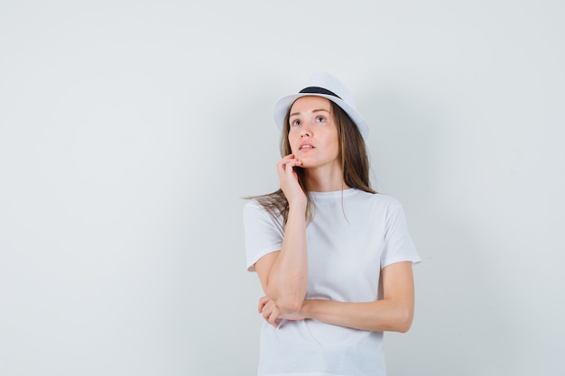 Mujer joven en camiseta blanca, sombrero mirando hacia arriba y mirando pensativo.