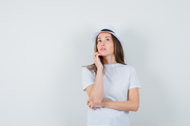 Mujer joven en camiseta blanca, sombrero mirando hacia arriba y mirando pensativo.