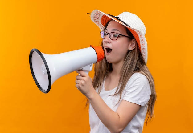 Mujer joven en camiseta blanca con sombrero de verano gritando al megáfono parado sobre la pared naranja