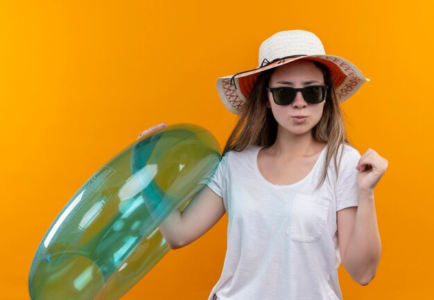 Mujer joven en camiseta blanca con sombrero de verano sosteniendo un anillo inflable apretando el puño mirando confiado con expresión seria de pie sobre la pared naranja