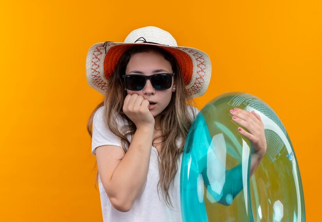 Mujer joven en camiseta blanca con sombrero de verano sosteniendo un anillo inflable mirando asombrado y sorprendido de pie sobre la pared naranja