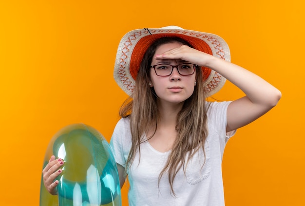 Mujer joven en camiseta blanca con sombrero de verano sosteniendo un anillo inflable mirando lejos con la mano sobre la cabeza para mirar a alguien o algo parado sobre una pared naranja