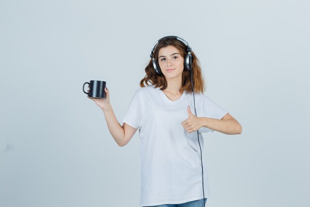 Mujer joven en camiseta blanca sosteniendo una taza de bebida, mostrando el pulgar hacia arriba y mirando alegre, vista frontal.