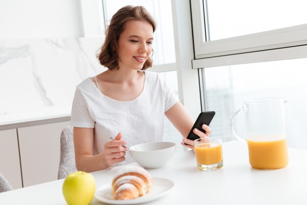 Mujer joven en camiseta blanca con teléfono móvil mientras desayuna en la cocina