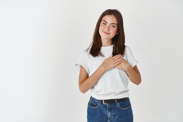 Mujer joven en camiseta casual cogidos de la mano en el corazón y sonriendo con cuidado y amor, de pie sobre la pared blanca