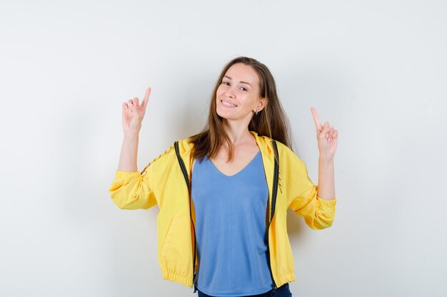 Mujer joven en camiseta, chaqueta apuntando hacia arriba y mirando confiado, vista frontal.