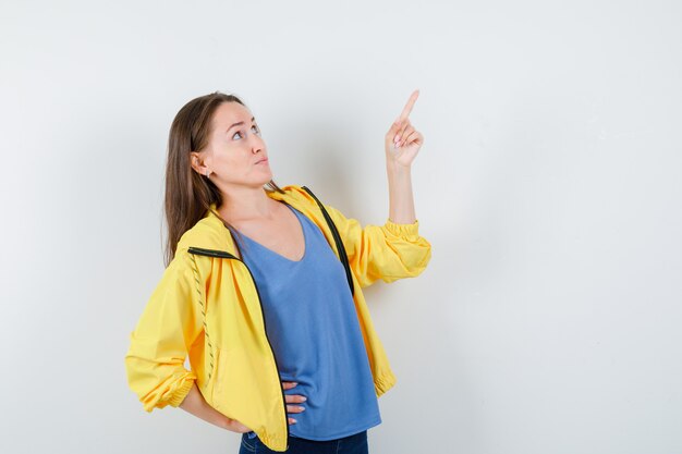 Mujer joven en camiseta, chaqueta apuntando hacia arriba y mirando enfocado, vista frontal.