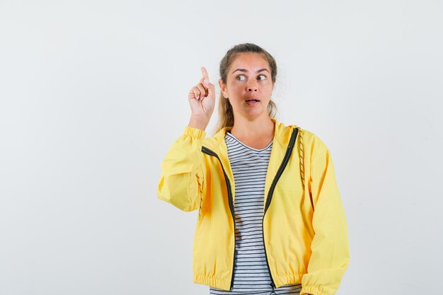 Mujer joven en camiseta, chaqueta apuntando hacia arriba y mirando pensativo, vista frontal.