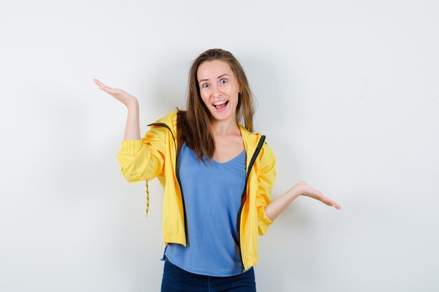 Mujer joven en camiseta, chaqueta haciendo gesto de escamas y mirando confiado, vista frontal.