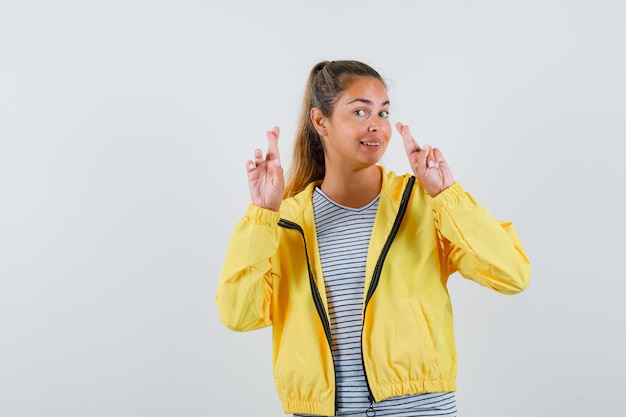 Mujer joven en camiseta, chaqueta manteniendo los dedos cruzados y mirando alegre, vista frontal.