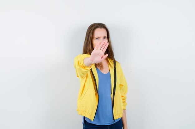 Mujer joven en camiseta, chaqueta mostrando gesto de parada y mirando molesto, vista frontal.