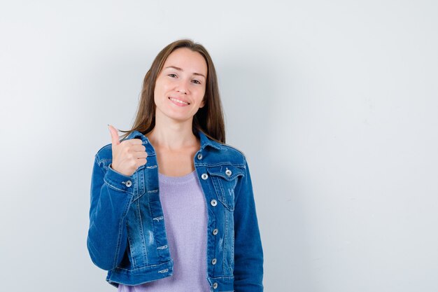 Mujer joven en camiseta, chaqueta mostrando el pulgar hacia arriba y mirando alegre, vista frontal.