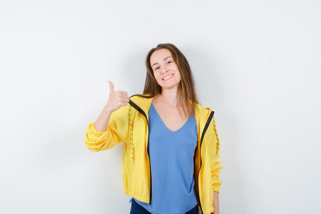 Mujer joven en camiseta, chaqueta mostrando el pulgar hacia arriba y mirando confiado, vista frontal.
