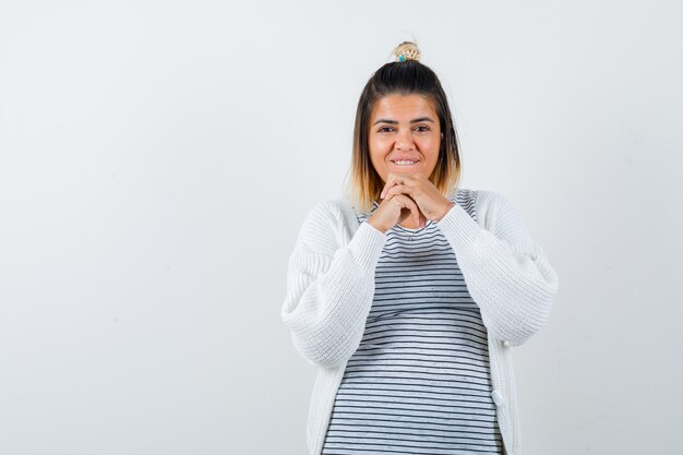 Mujer joven en camiseta, chaqueta de punto manteniendo las manos juntas sobre el pecho y mirando positivo, vista frontal.