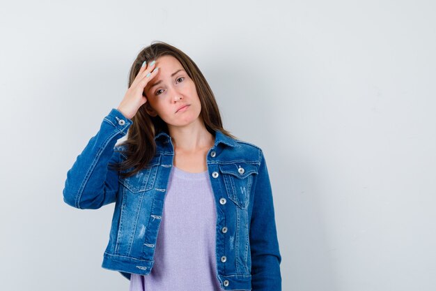 Mujer joven en camiseta, chaqueta que sufre de dolor de cabeza y mira angustiada, vista frontal.