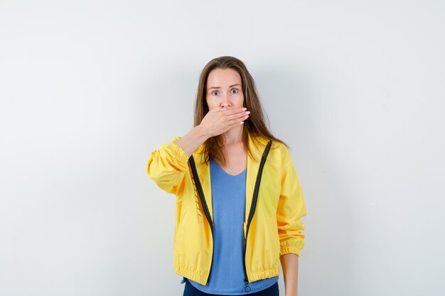 Mujer joven en camiseta, chaqueta sosteniendo la mano en la boca y mirando sorprendido, vista frontal.