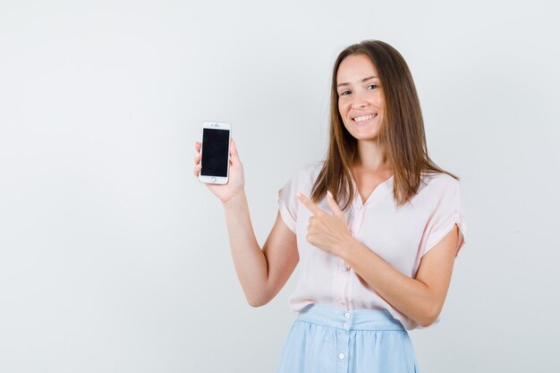 Mujer joven en camiseta, falda apuntando al teléfono móvil y mirando alegre, vista frontal.