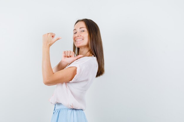 Mujer joven en camiseta, falda apuntando hacia atrás con los pulgares y mirando alegre.