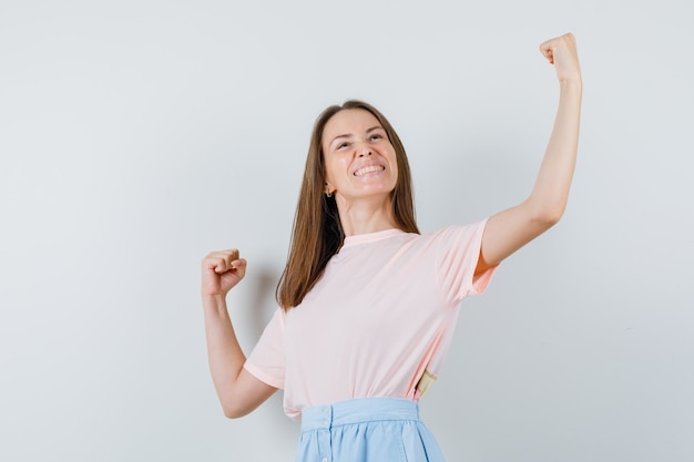 Mujer joven en camiseta, falda mostrando gesto de ganador y mirando feliz, vista frontal.