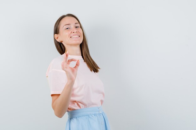 Mujer joven en camiseta, falda mostrando gesto ok y mirando complacido, vista frontal.
