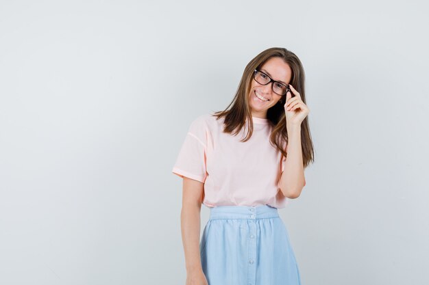 Mujer joven en camiseta, falda sosteniendo los dedos en las gafas y mirando alegre, vista frontal.