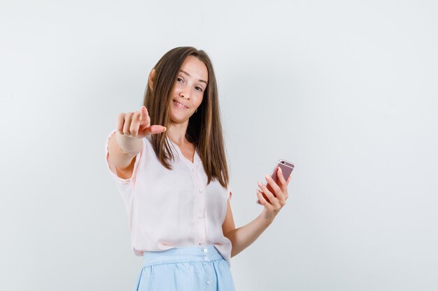 Mujer joven en camiseta, falda sosteniendo teléfono móvil y apuntando a cámara, vista frontal.