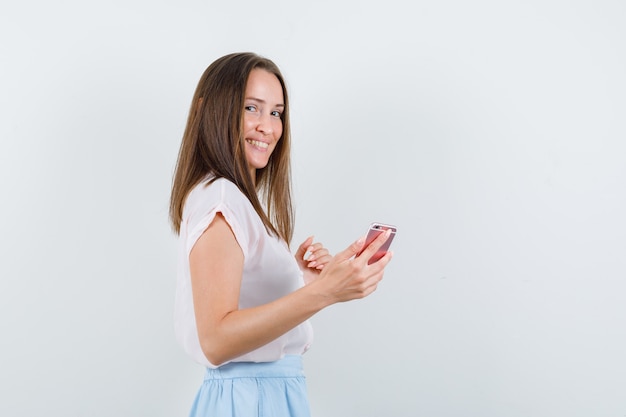 Mujer joven en camiseta, falda sosteniendo teléfono móvil y mirando contento.