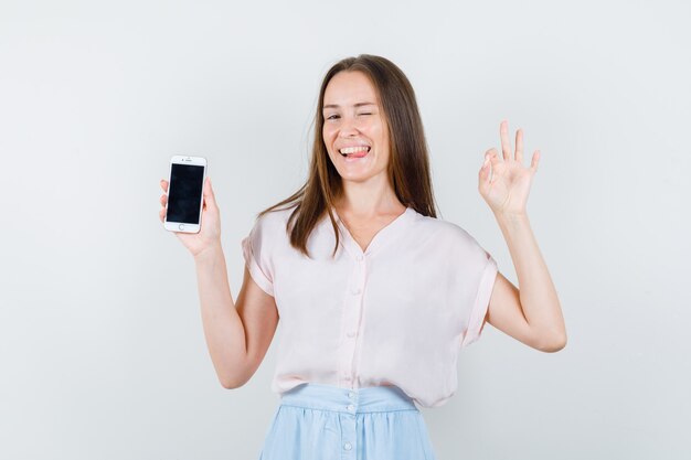 Mujer joven en camiseta, falda sosteniendo teléfono móvil con signo ok y mirando divertido, vista frontal.