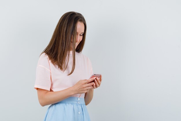 Mujer joven en camiseta, falda usando teléfono móvil y mirando feliz, vista frontal.