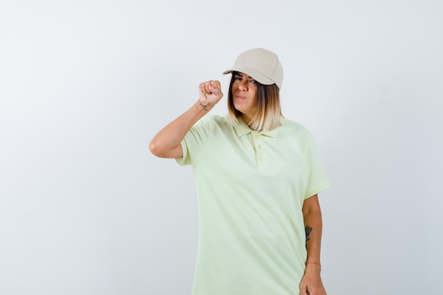 Mujer joven en camiseta, gorra levantando el puño cerca de la cara y mirando serio, vista frontal.