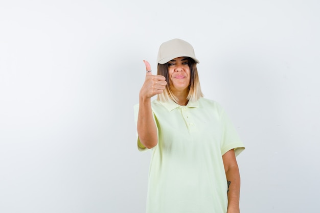 Mujer joven en camiseta, gorra mostrando el pulgar hacia arriba y mirando alegre, vista frontal.