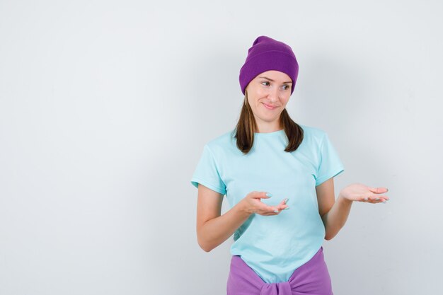 Mujer joven en camiseta, gorro fingiendo mostrar algo y mirando alegre, vista frontal.