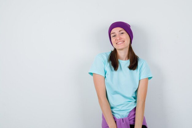 Foto gratuita mujer joven en camiseta, gorro posando mientras está de pie y mirando alegre, vista frontal.