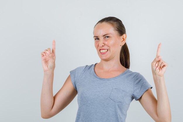 Mujer joven en camiseta gris apuntando hacia arriba y apretando los dientes, vista frontal.