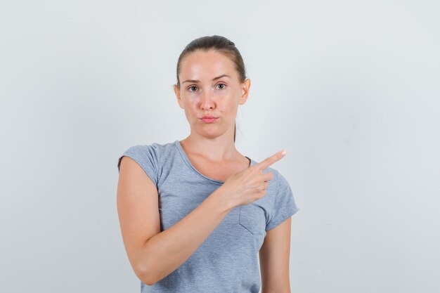 Mujer joven en camiseta gris apuntando hacia un lado y mirando serio, vista frontal.