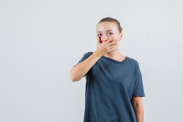 Mujer joven en camiseta gris cubriendo la boca con la mano y mirando sorprendido