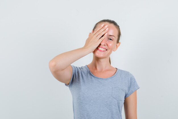 Mujer joven en camiseta gris cubriendo un ojo con la mano y mirando divertido, vista frontal.