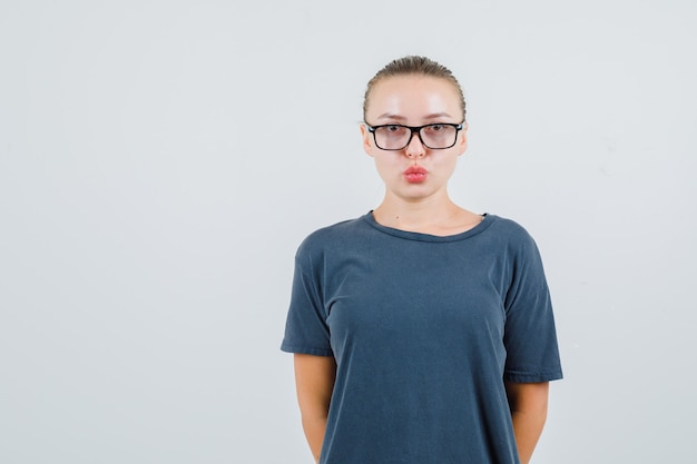 Mujer joven en camiseta gris, gafas manteniendo los labios doblados y mirando inteligente