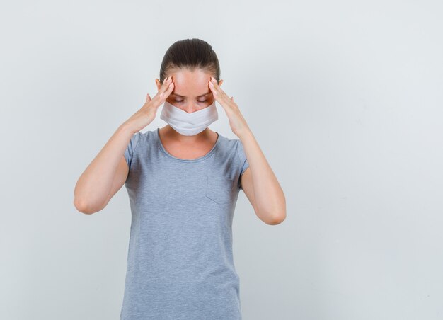 Mujer joven en camiseta gris, máscara sosteniendo los dedos en la frente y mirando exhausto, vista frontal.