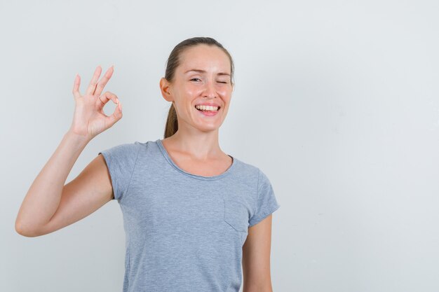 Mujer joven en camiseta gris mostrando gesto ok y guiño de ojo, vista frontal.