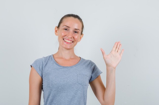 Mujer joven en camiseta gris mostrando la palma y mirando alegre, vista frontal.