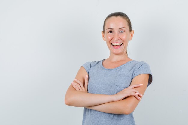 Mujer joven en camiseta gris de pie con los brazos cruzados y mirando alegre, vista frontal.