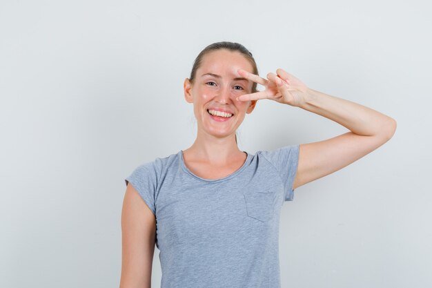 Mujer joven en camiseta gris que muestra el signo v cerca del ojo y luciendo alegre, vista frontal.