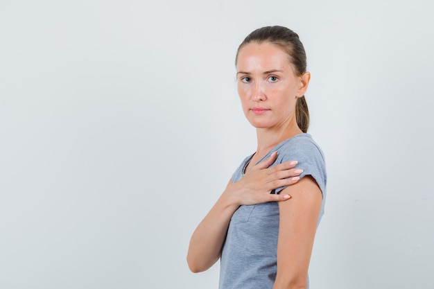Mujer joven en camiseta gris sosteniendo la mano cerca del corazón y mirando seria, vista frontal.
