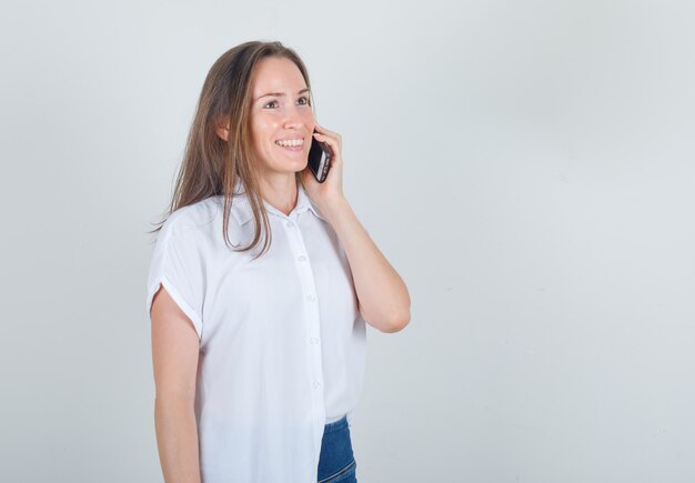 Mujer joven en camiseta, jeans hablando por teléfono inteligente y mirando alegre