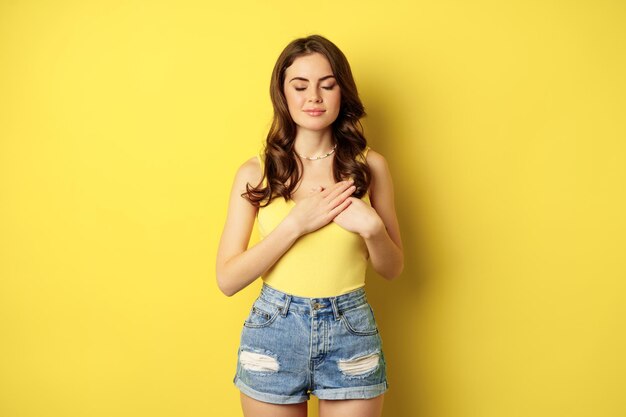 Mujer joven en camiseta sin mangas, tomándose de la mano en el corazón con cuidado y ternura, sonriendo a la cámara, luciendo agradecida, agradecida, de pie sobre un fondo amarillo.