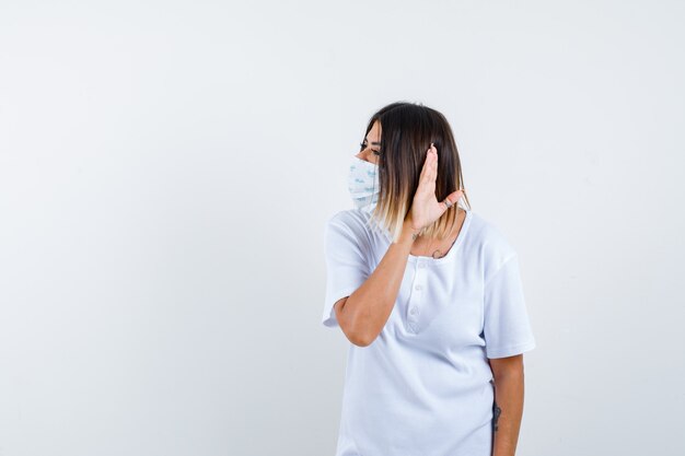 Mujer joven en camiseta, máscara sosteniendo la mano detrás de la oreja y mirando sorprendido, vista frontal.