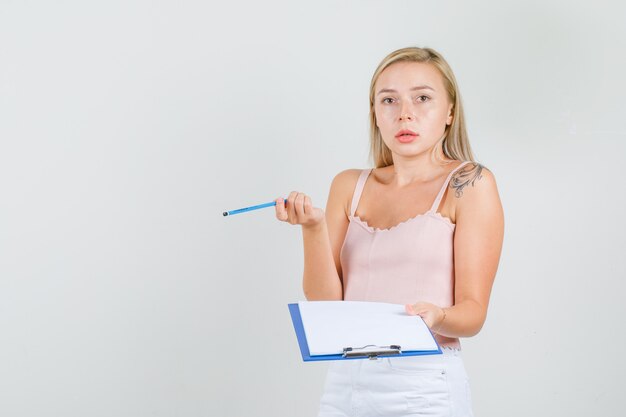 Mujer joven en camiseta, mini falda sosteniendo lápiz y portapapeles y mirando confundido