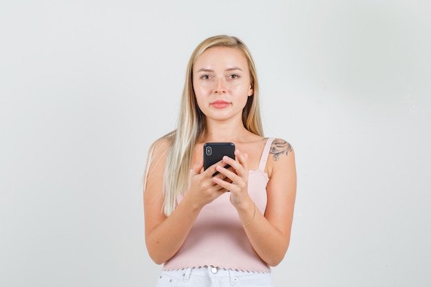 Mujer joven en camiseta, mini falda sosteniendo smartphone y mirando a cámara