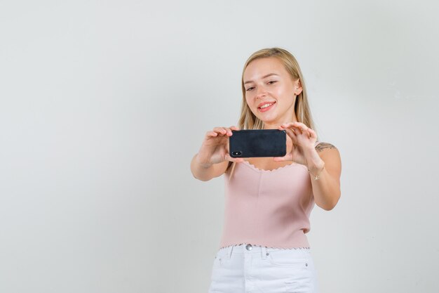 Mujer joven en camiseta, mini falda tomando fotos en el teléfono y sonriendo